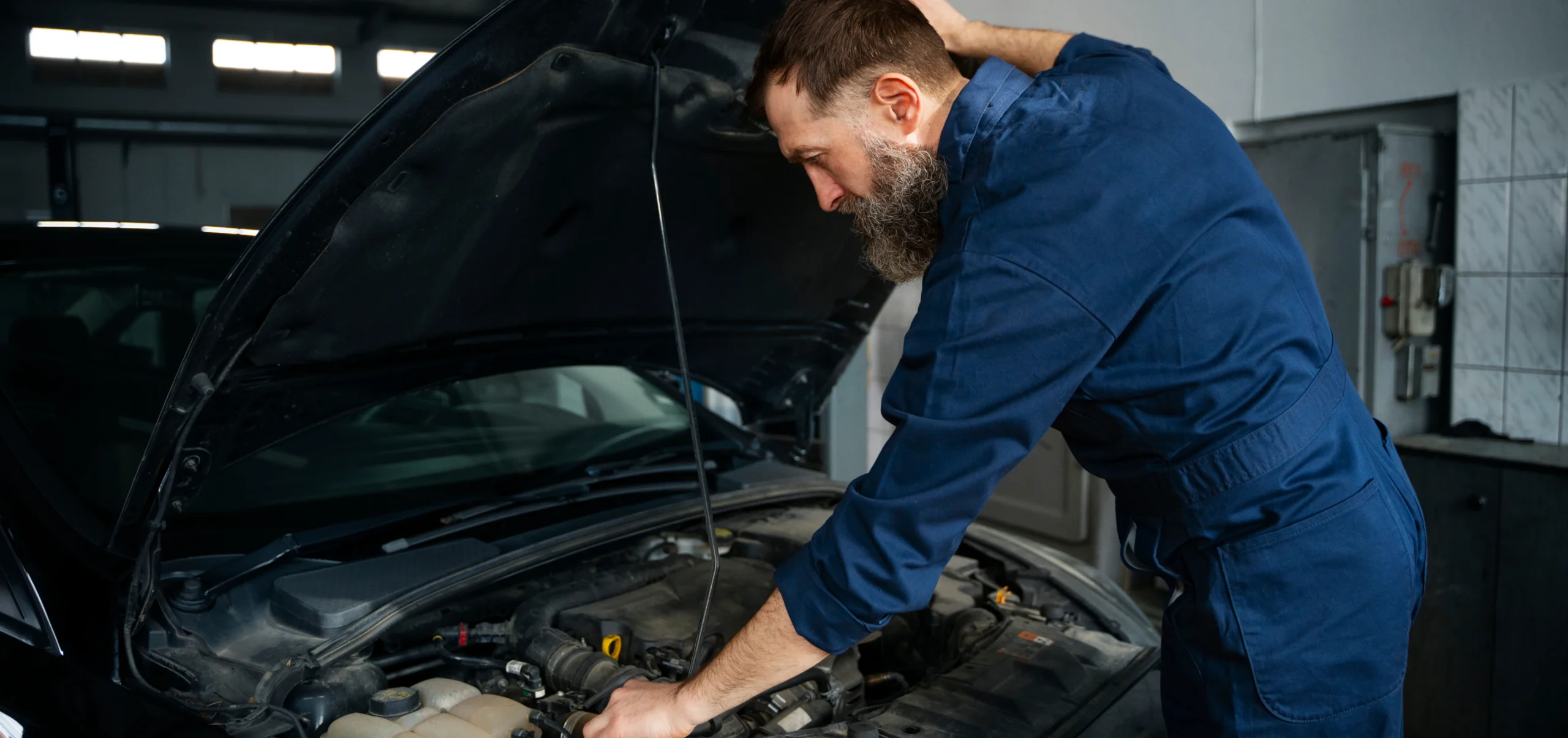 A man with a beard in blue overalls repairs a car in an auto shop in Houston.