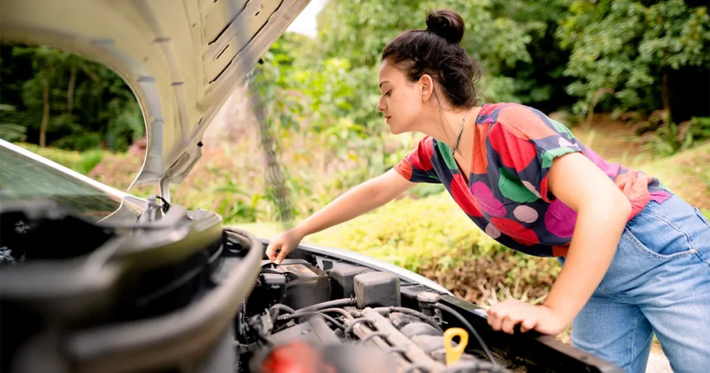 A woman leaning over an open car hood in a driveway, inspecting the engine for leaks or smoke.