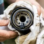 A person holding a dirty, used oil filter wrapped in a grease-stained white cloth to show carbon buildup.