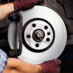 A mechanic wearing red gloves holding a clean brake rotor disc during a brake system repair.
