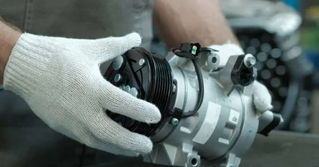 A technician wearing white work gloves holding a new silver automotive AC compressor unit.