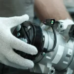 A technician wearing white work gloves holding a new silver automotive AC compressor unit.