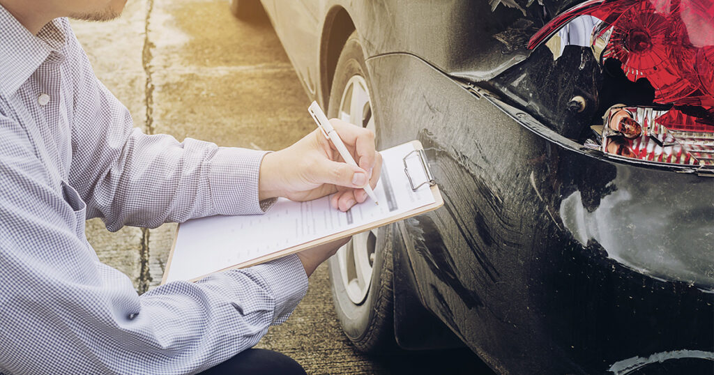 An investigator writes on a clipboard while inspecting a car's smashed taillight and bumper scratches after an accident.