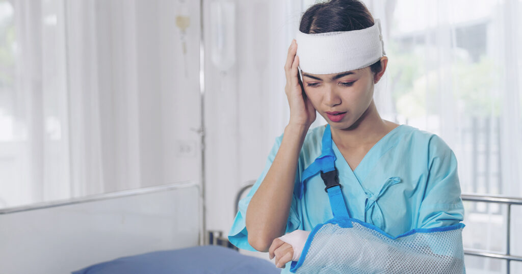 A woman in a hospital gown with a head bandage and arm sling holds her temple, appearing in pain after a brain injury.