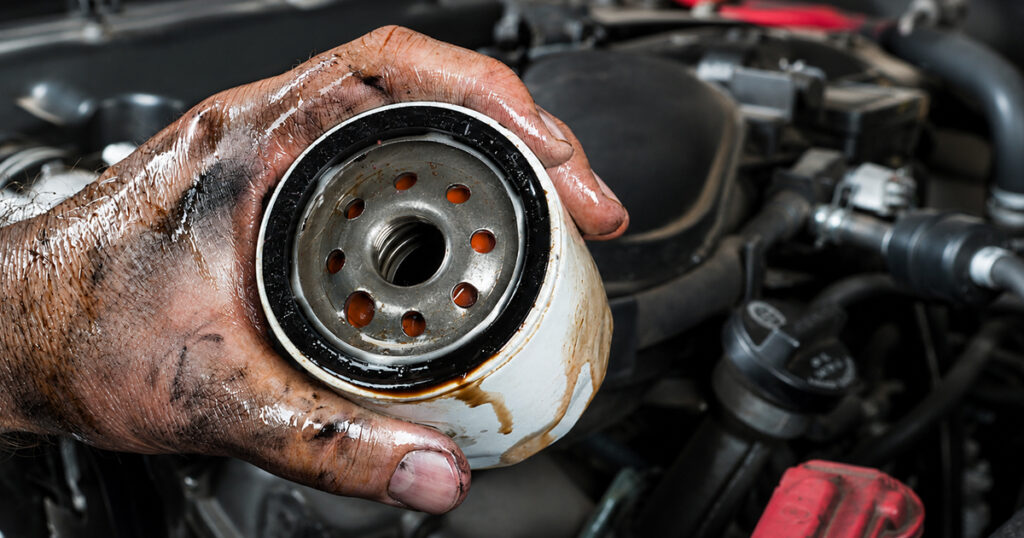 Greasy hand holding a used oil filter over a car engine.
