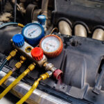 A close-up view of a car's engine bay with an AC manifold gauge set resting on the front plastic cover. The diagnostic tool features prominent blue and red pressure dials connected to thick yellow and blue hoses, typically used for servicing the vehicle's air conditioning system.