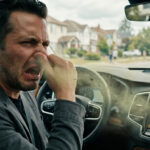 A man sitting in the driver's seat of a car, pinching his nose in disgust as visibly dirty, greenish air blows out of the dashboard vents