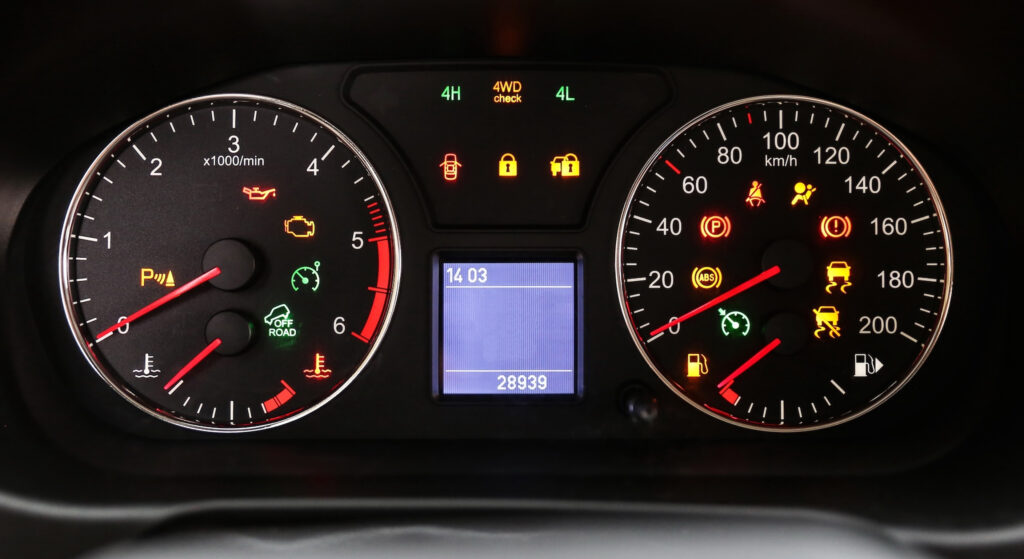 A close-up view of a car's instrument cluster with numerous red, yellow, and green warning lights illuminated. The dashboard features an analog tachometer on the left, an analog speedometer on the right, and a small central digital display showing numbers.