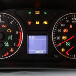 A close-up view of a car's instrument cluster with numerous red, yellow, and green warning lights illuminated. The dashboard features an analog tachometer on the left, an analog speedometer on the right, and a small central digital display showing numbers.