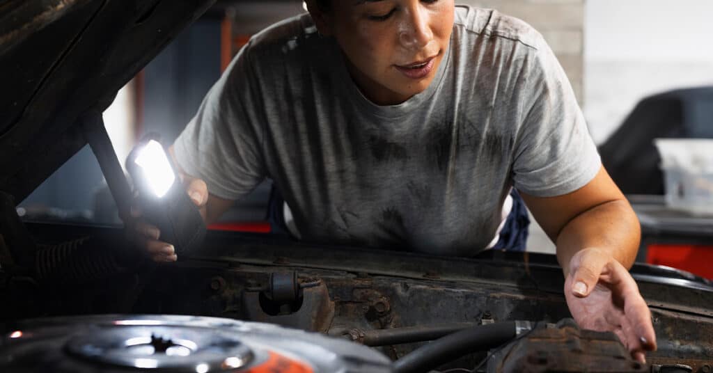 A mechanic uses a flashlight to inspect an engine under the hood of a car.