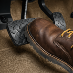 Brown leather boot pressing a car pedal on a beige floor mat.