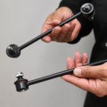 Person in a black shirt holding two sway bar links against a plain background.