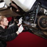 A mechanic in a black uniform and work gloves inspects the suspension and rear undercarriage of a car raised on a lift against a deep red background.