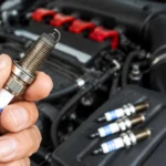 Hand holding a spark plug beside a car engine.