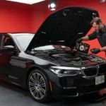 A mechanic in a gray uniform is pouring motor oil into a black BMW sedan inside a red-walled garage.
