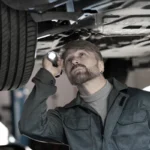 Mechanic using flashlight to inspect car underside on vehicle lift.