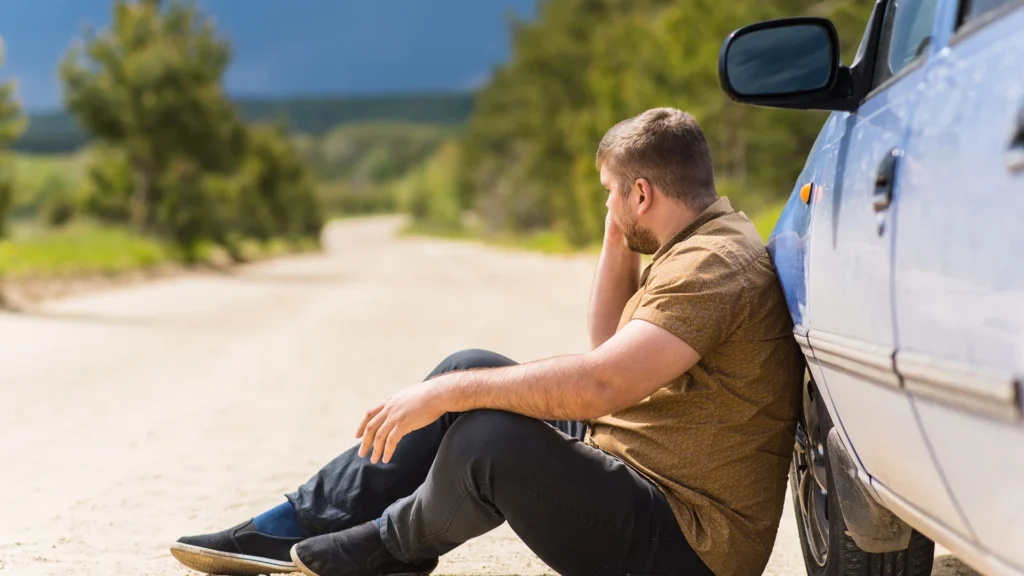 A stranded driver sitting on a dirt road leaning against his car while making a phone call