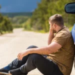 A stranded driver sitting on a dirt road leaning against his car while making a phone call