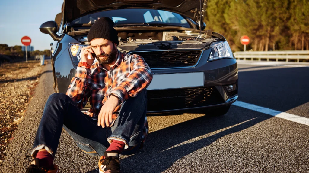 A stranded man sitting on the asphalt on a smartphone in front of a car with an open hood