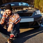 A stranded man sitting on the asphalt on a smartphone in front of a car with an open hood